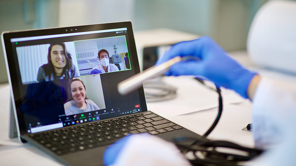A professor teaches a class over a Zoom call in the lab with two students on the screen. 