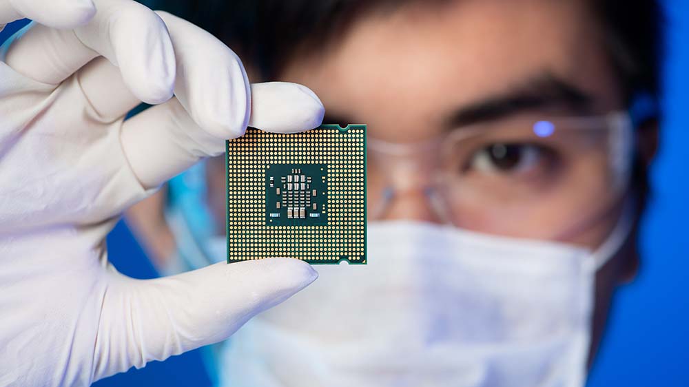 A researcher, slightly out of focus, holds up a small computer chip to the camera.
