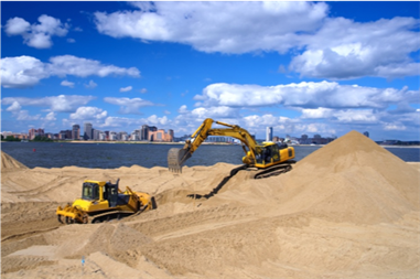 A yellow bull dozer and crane are on the beach gathering and moving sand around on a beach. A large body of water and a city are in the background.