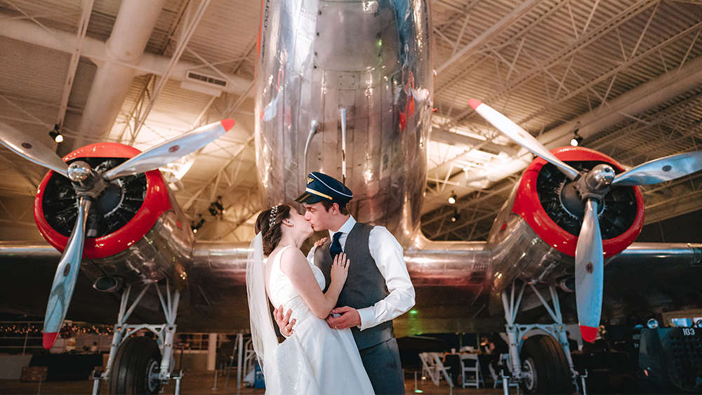 Katie and Steven, former aerospace engineering students, are dressed in their bride and groom wedding attire. Katie is wearing a white bridal gown with veil and Steven is wearing a dark gray vest, white collared shirt and navy blue tie along with a navy blue and gold pilots hat. Steven is holding Katie in his arms as her kisses her under the nose of a restored DC-3 aircraft of silver and red color.