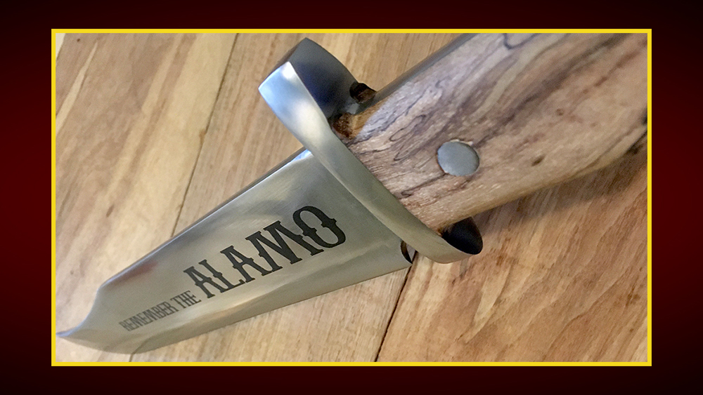 A bowie knife with its sharp edge pointing away. It has a wooden handle and the words "Remember the Alamo" etched into the blade.