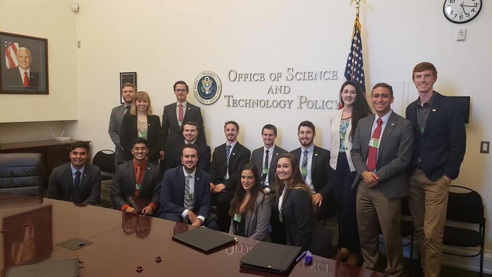 A group of students in business professional attire stand in the Office of Science and Technology Policy