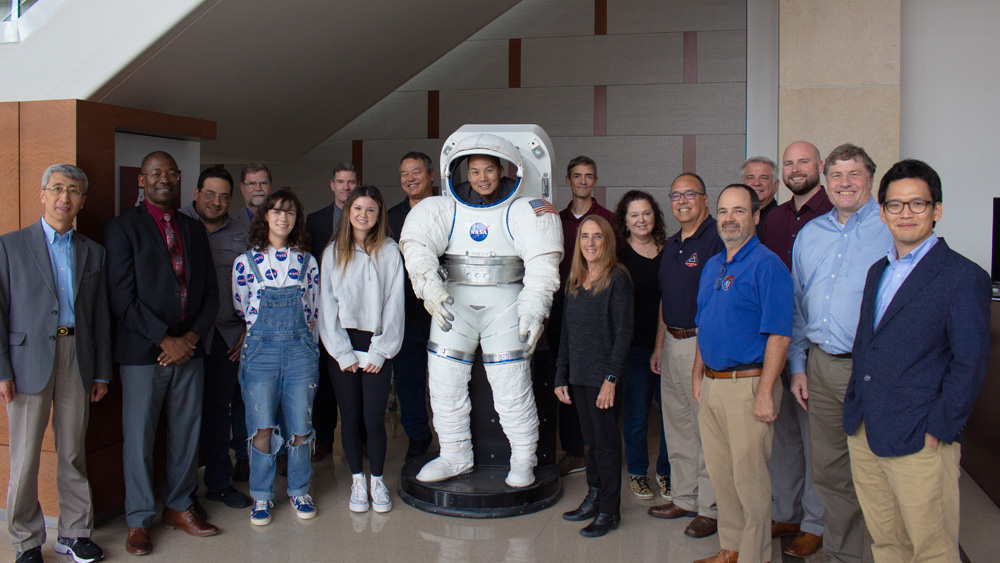 A group of NASA representatives, their families and Texas A&M faculty stand together around a life-sized space suit model and smile at the camera.