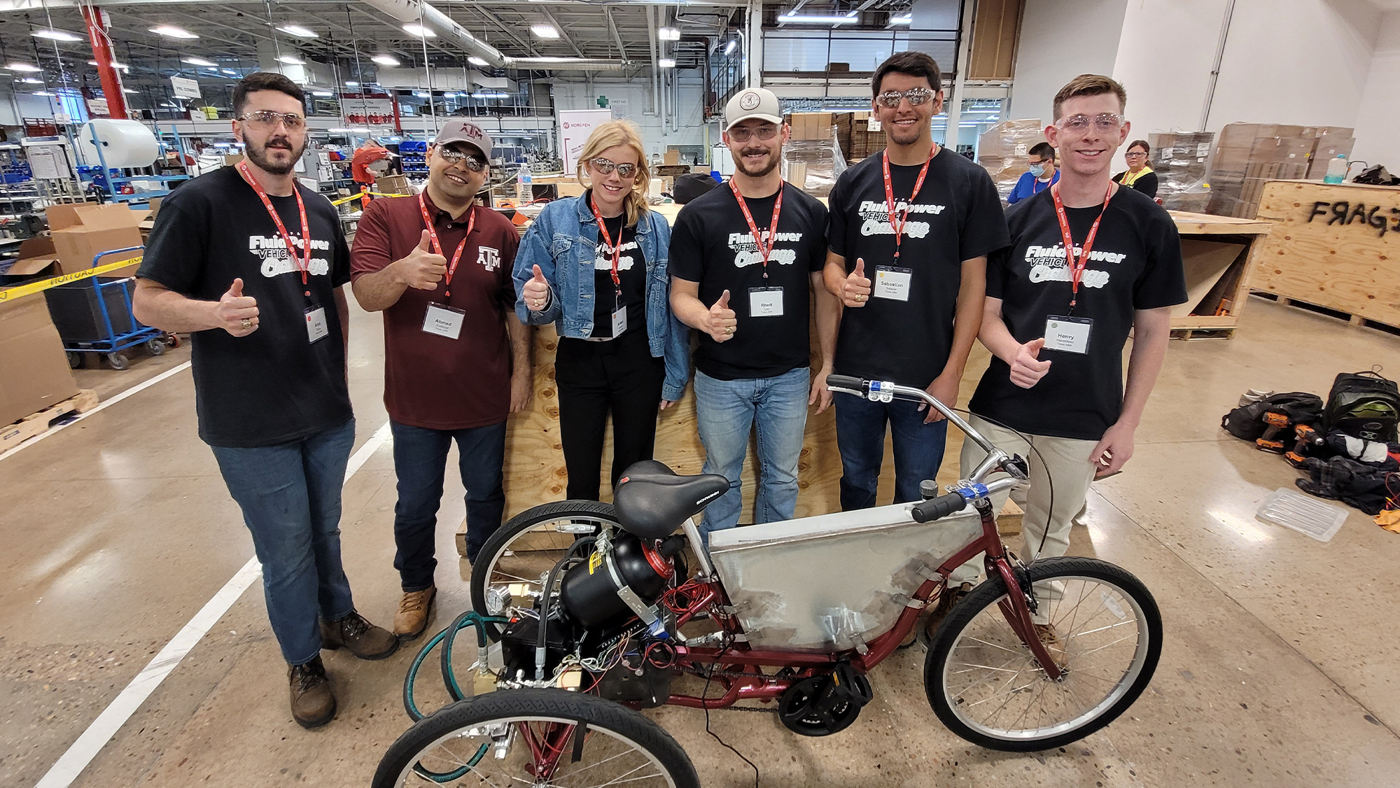 Inside the Norgren facility, the Aggie student team and faculty supervisor Dr. Ahmed Abdelaal give the thumbs-up sign. In the foreground is the tricycle the team designed for the Fluid Power Vehicle Challenge. 