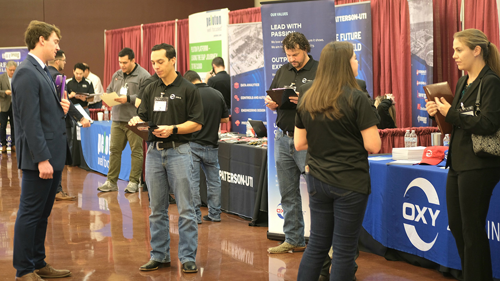 Row of tables sporting energy company logos and banners line a wall behind company representatives and students speaking to each other in one-on-one conversations at a career fair.