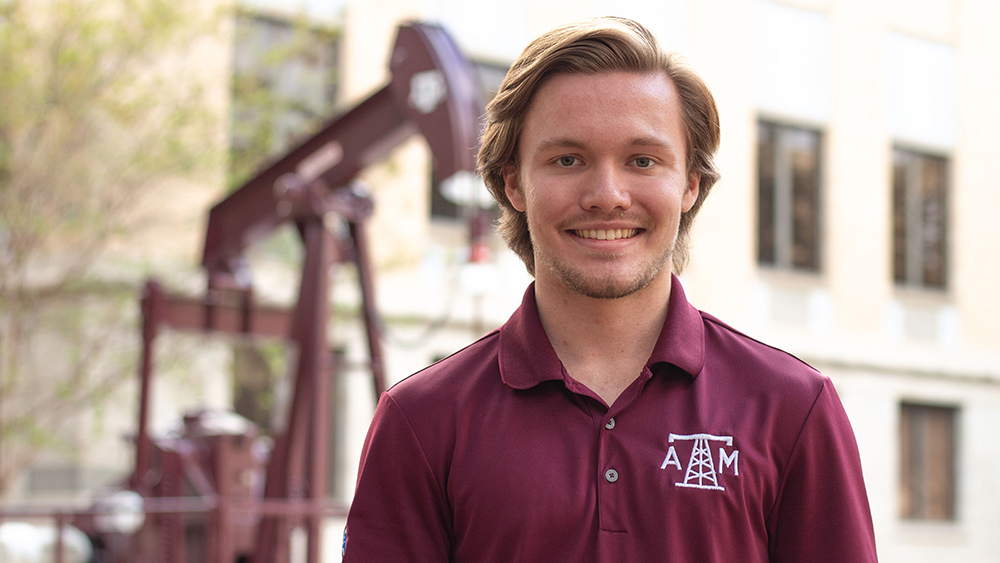 Nathan Hazlett standing outdoors in front of the Aggie Well No. 1 pump jack on the Texas A&M University campus