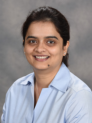 Dr. Farheen Naqvi wearing a blue collared shirt in front of a grey patterned background