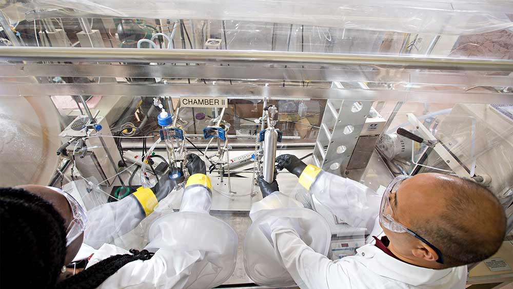 Female and male researchers wearing lab coats and other safety gear while working with an enclosed experiment station