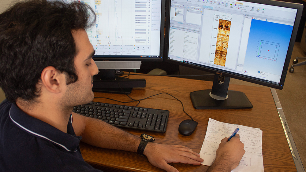 male petroleum engineering student seated at a desk and writing equations with a pencil while reviewing a borehole graphic showing vertical images of color-coded data changes on a computer monitor