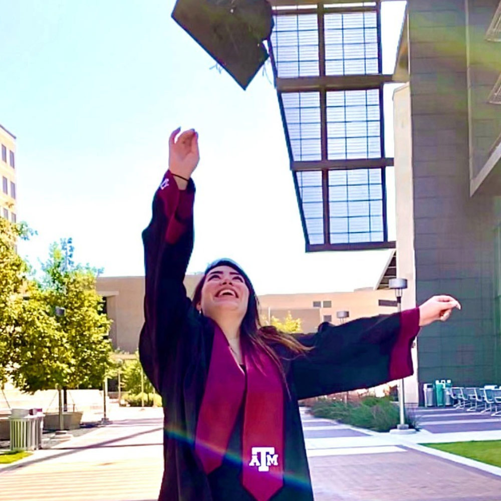Student wearing her Texas A&amp;M cap and gown. She is throwing her cap into the air outside the Zachary building. 