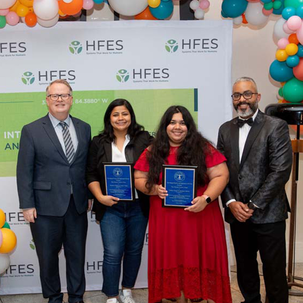 Dr. Ranjana Mehta and Oshin Tyagi hold plaques and stand between two men in front of a colorful balloon arch and a Human Factors and Ergonomics Society banner