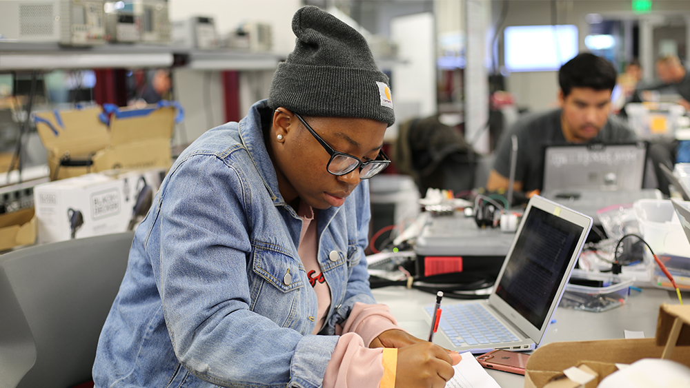 A female wearing glasses and a hat takes notes inside the Fischer Engineering Design Center