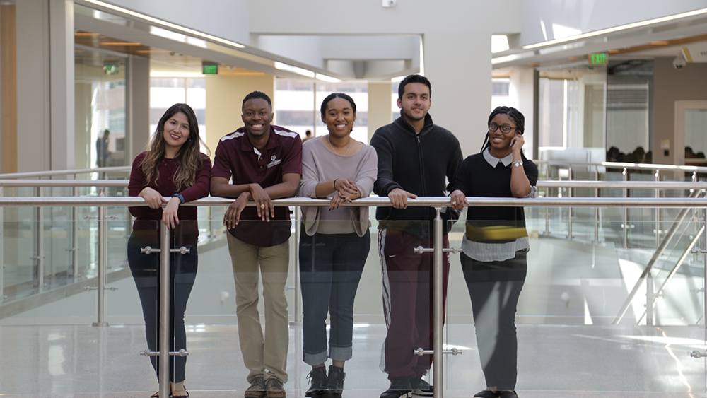 Five students pose for a photo, leaning over the railing in the ZACH building