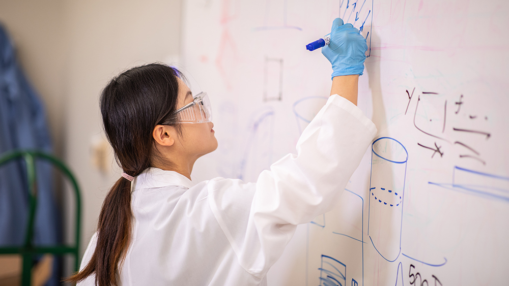 A female student wearing a white lab coat, safety goggles and blue gloves writes on a white board with a blue marker.