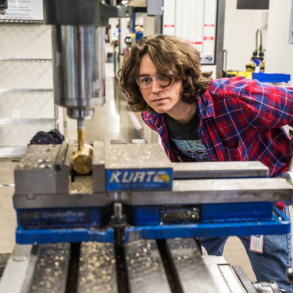 student wearing protective eyewear intently watching a drill press in a workshop environment.