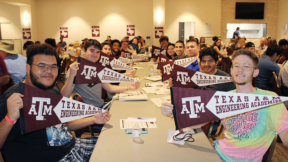 A group of diverse students sitting at a table, smiling and holding Texas A&amp;M Engineering Academies maroon pennants. The room is full of students celebrating their involvement in the Engineering Academies program.