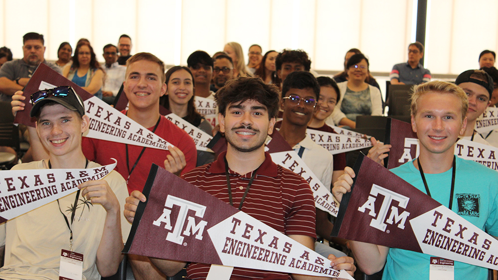 Several students sitting together in a classroom, proudly holding maroon Texas A&amp;M Engineering Academies pennants. A large group of peers is visible in the background, smiling and participating.