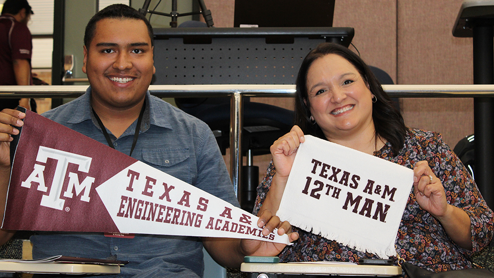 A young man and woman sitting close together, smiling brightly and holding Texas A&amp;M themed items—a maroon pennant and a white "12th Man" rally towel.