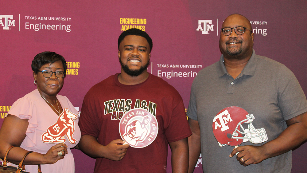 A family of three standing in front of a Texas A&amp;M Engineering Academies backdrop. The young man wears a maroon Texas A&amp;M shirt, while his parents hold Texas A&amp;M themed cutouts, including a football helmet.