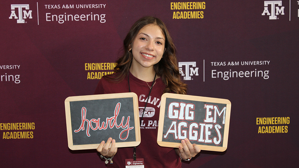 A young woman posing in front of a maroon Texas A&amp;M Engineering Academies backdrop, holding two chalkboard signs that say "Howdy" and "Gig 'Em Aggies."