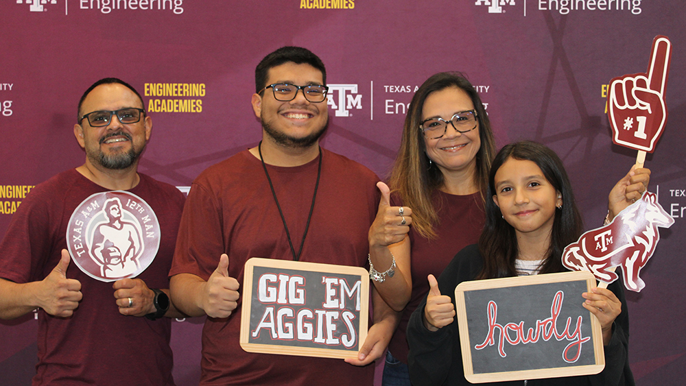 A family of four standing together in front of the Texas A&amp;M Engineering Academies backdrop. They are all wearing maroon and holding Texas A&amp;M props, including chalkboard signs saying "Howdy" and "Gig 'Em Aggies," a foam finger, and a 12th Man logo cutout.