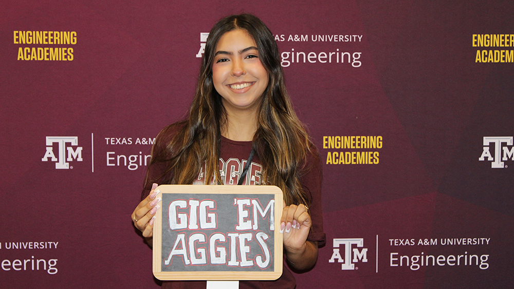 A young woman smiling in front of the Texas A&amp;M Engineering Academies backdrop, proudly holding a chalkboard sign that reads "Gig 'Em Aggies."