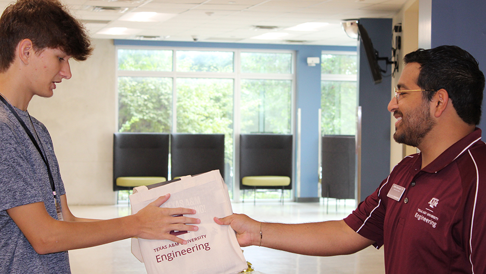 Hispanic male wearing a maroon polo smiles while handing a back of papers across a table to a male wearing a grey t-shirt. 