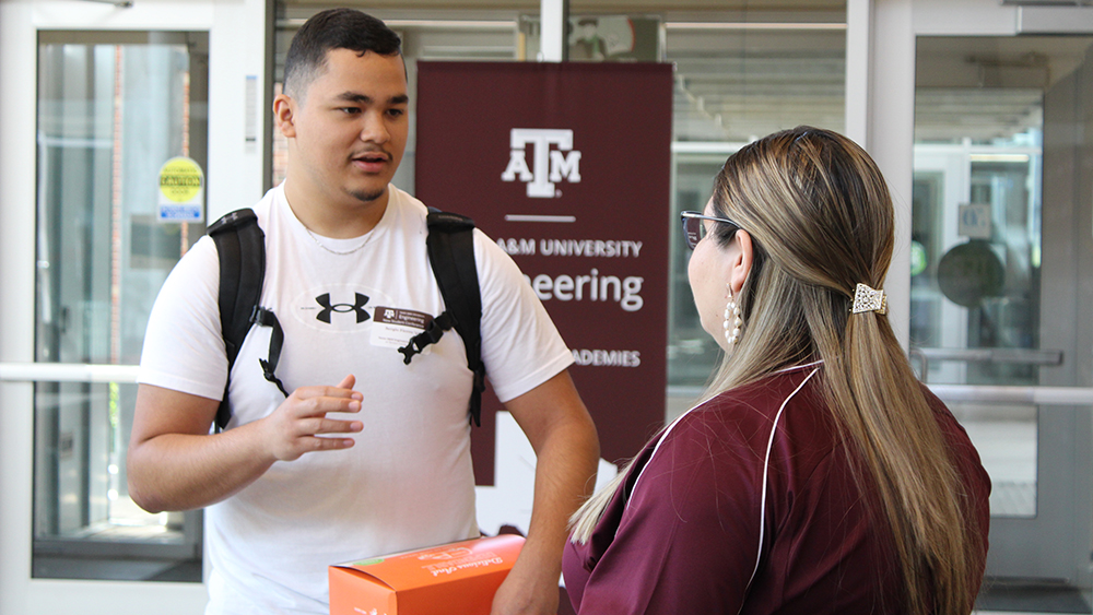 Male student in white shirt stands talking to a female Engineering Academy staff member in maroon shirt whose back is to the camera. 