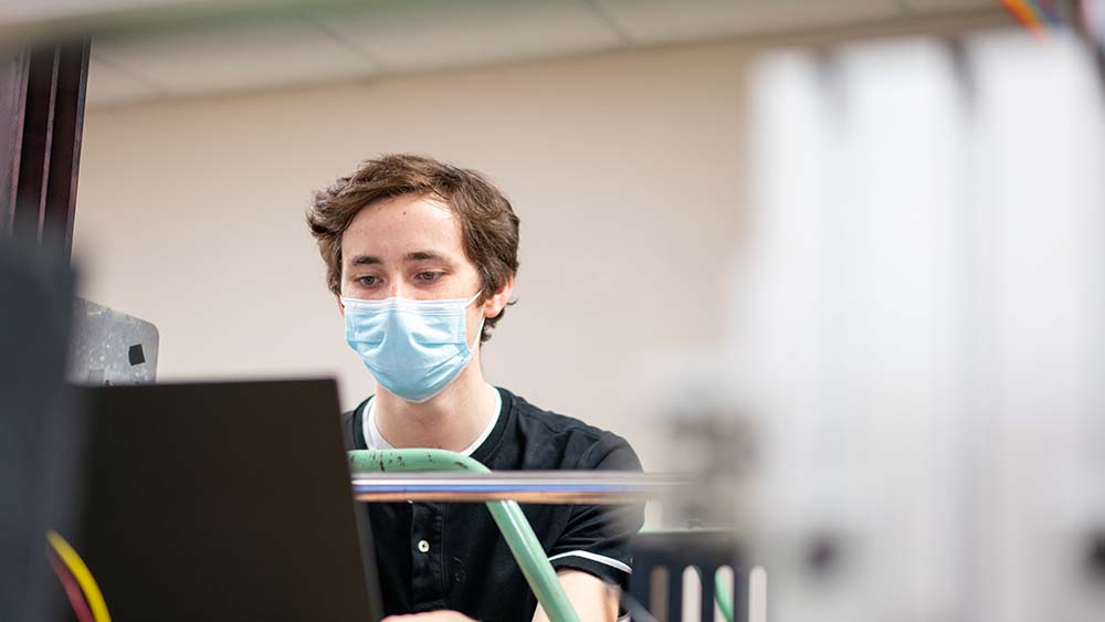 A man sits in a lab, wearing a blue mask on his face.