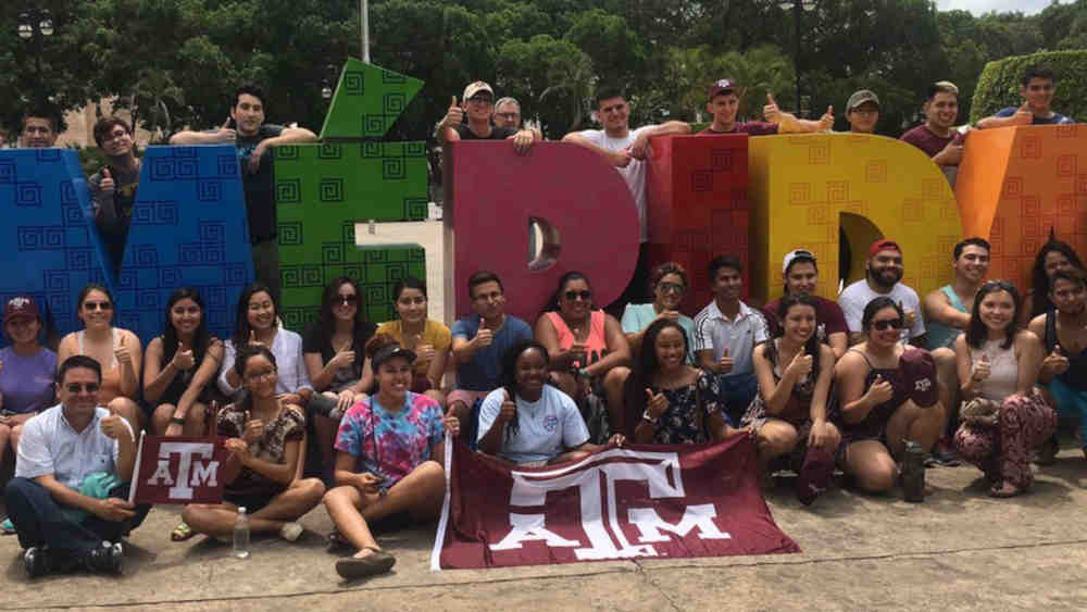 Students standing and sitting around the Merida sign with TAMU flag in front
