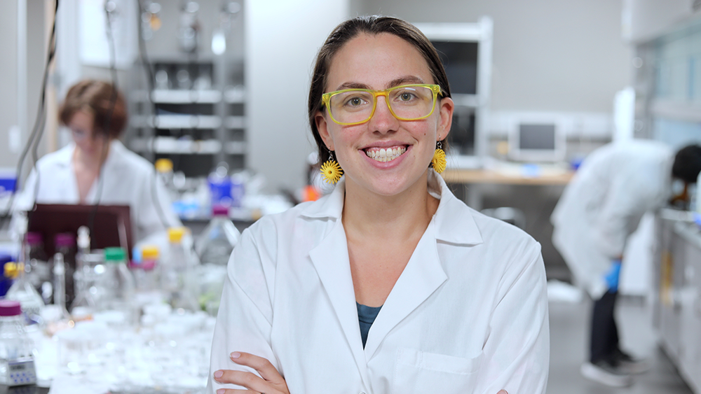 A woman wearing a white lab coat and yellow glasses smiles with her arms crossed, standing in a lab