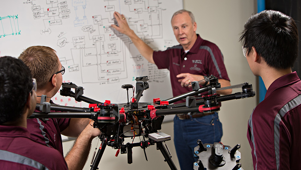 Male instructor pointing to a white board with diagrams. Three male students seated around a drone looking toward him.
