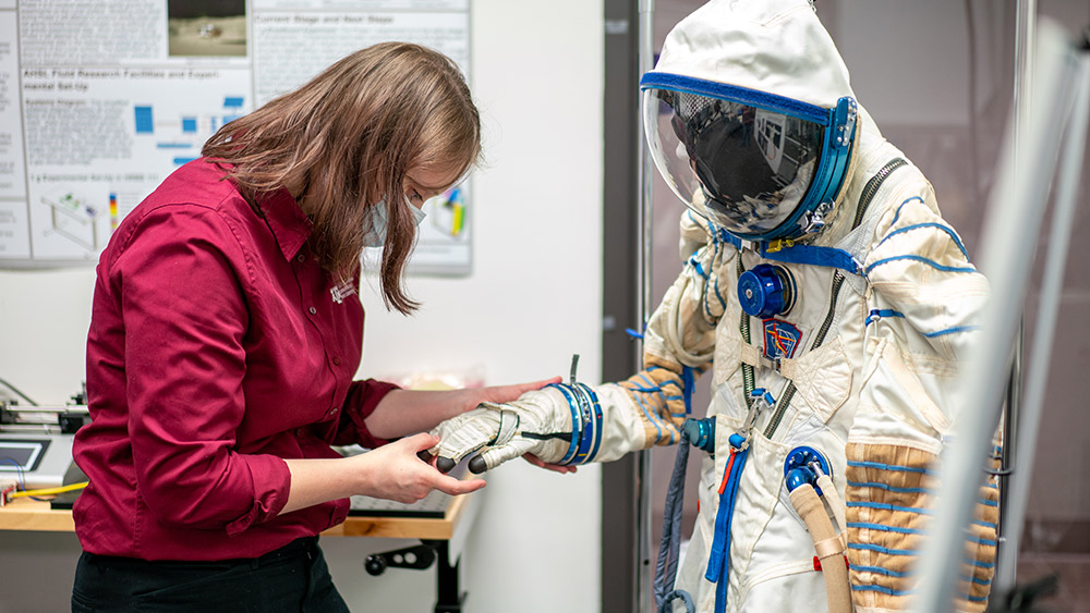 Female student standing in front of an astronaut suit, holding and looking down at the suit's glove.