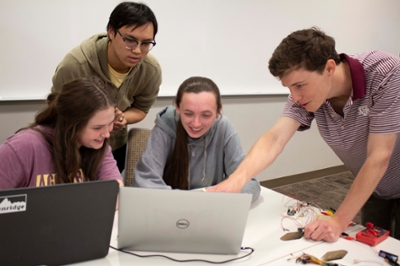 Two female and two male students look at a computer screen, one of the men is pointing at it.