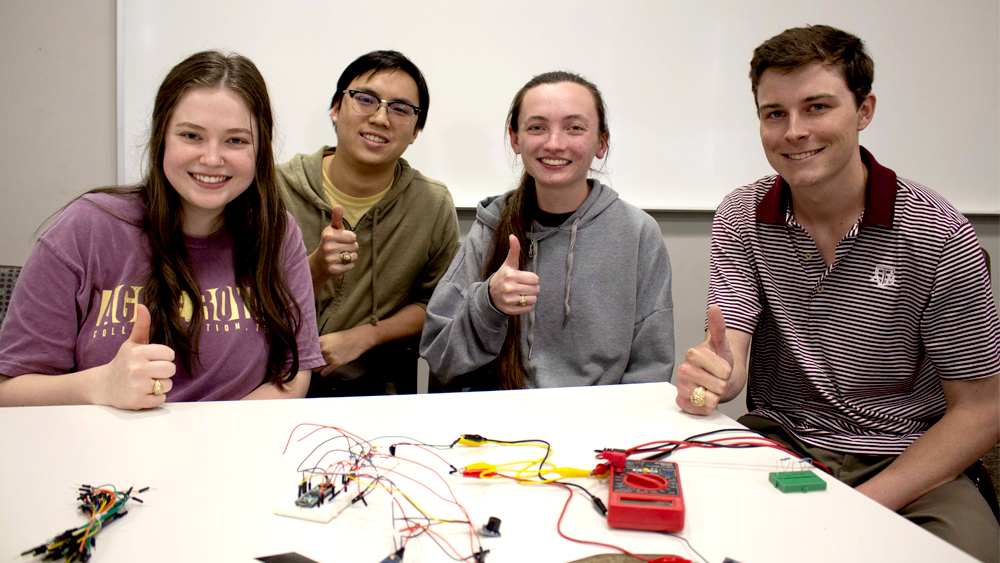 Four students, two male and two female, sit at a design table. They are smiling at the camera and giving the thumbs up gig 'em sign.