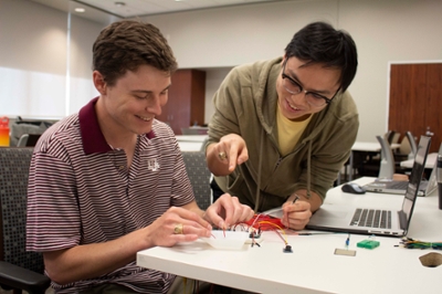 Two male students work at a design table. They are working with a series of wires and a container of liquid to test the electric connection of their circuit.