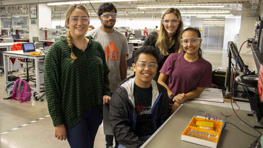 Five students, three female and two male, are in a design center. They are smiling at the camera, all wearing safety goggles.