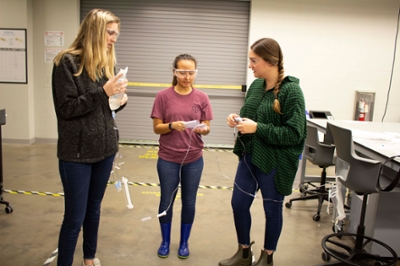 Three female students stand in a design studio. They are holding the tubing of an IV bag between them and are talking.