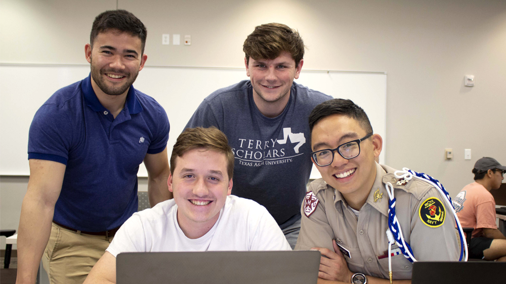 Five male students gathered around a laptop, smiling at the camera.