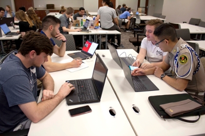 Four male students sit at a table, all working on their personal laptops. Around them are other tables also with students working.