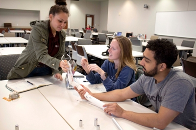 Three students, two female and one male, sit at a design table. They are holding a plastic and PVC pipe prototype of a bendable clamp holder.