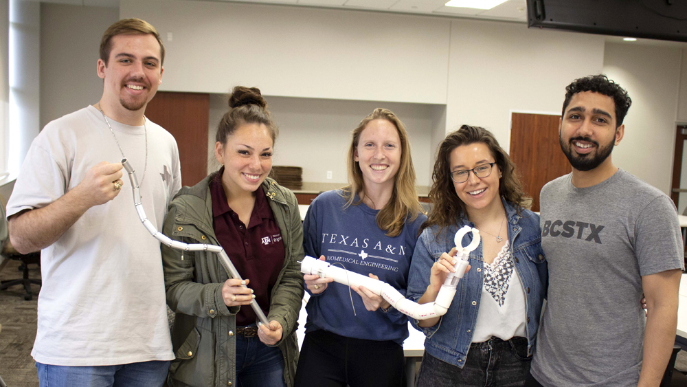 A group of five students, two men and three women, stand smiling at the camera. They are holding a plastic and PVC pipe version of a their flexible clamp holder.