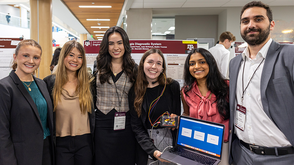 Six smiling students, with one wearing a device in her ear connected by wires to the laptop she's holding and the small box-like device held by another student.