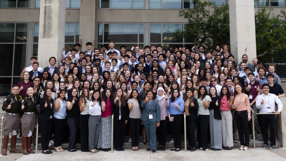 A crowd of students in professional dress stands in front of a large campus building.