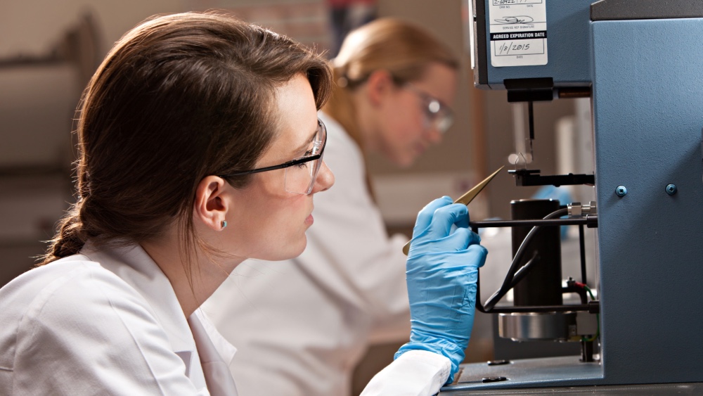 A young woman in lab coat, glasses, and medical gloves adjusts a sample in a piece of laboratory equipment using a pair of long tweezers.