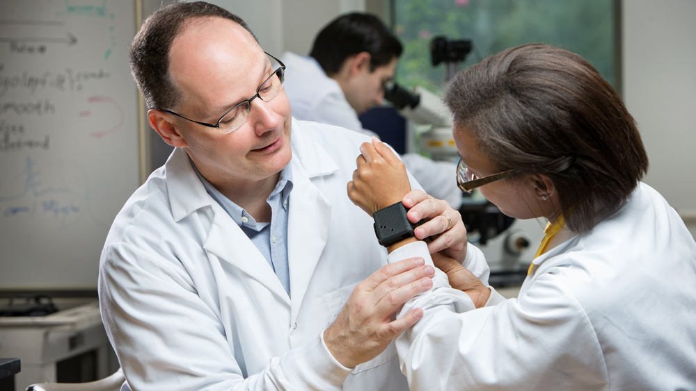 Two researchers in lab coats inspect a square-shaped medical device worn on one researcher’s wrist.