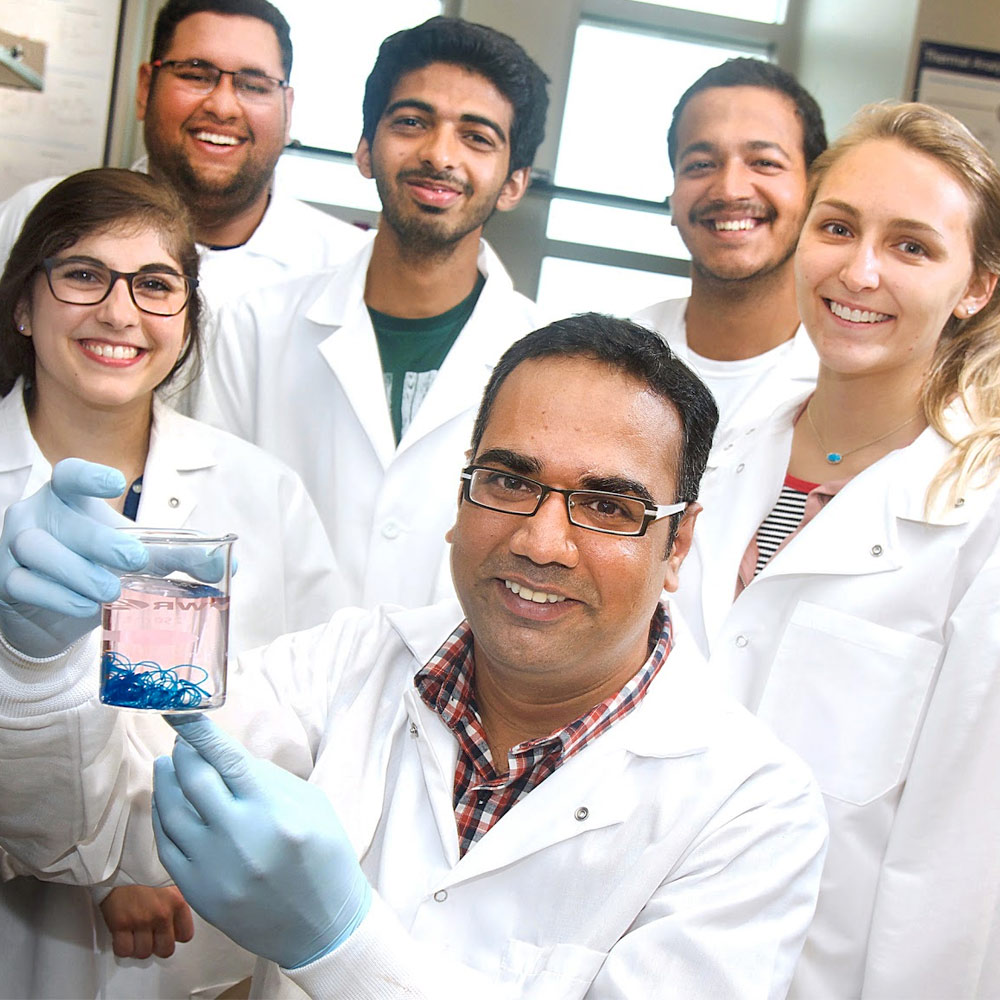 Professor holding up beaker in biomedical lab surrounded by students in white coats smiling