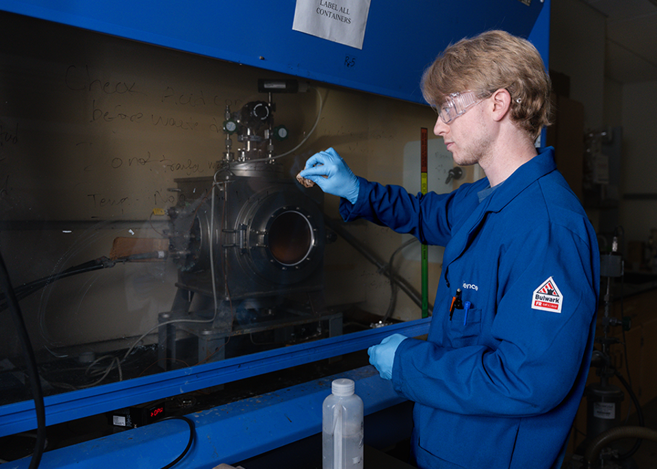 A technician in a blue lab coat conducts an experiment at a laboratory workstation, using gloves to handle equipment while focusing on a device. 