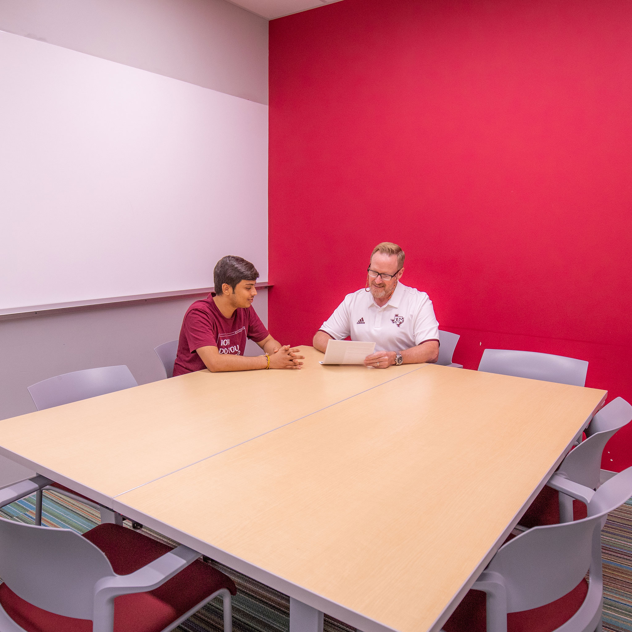 Two men discuss some paperwork at a table in a small conference room.