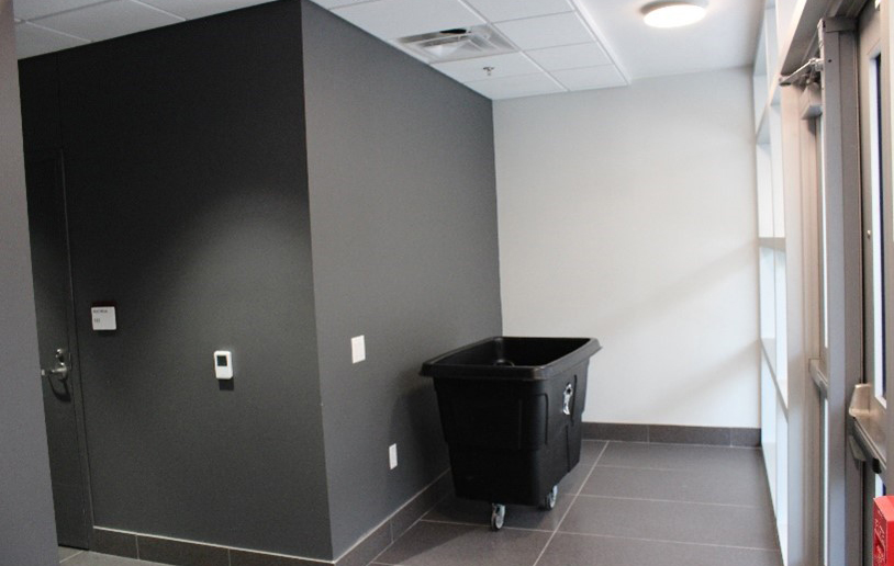 Large wheeled black trash bin in corner of hallway.